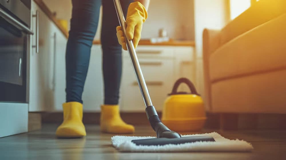 Person mopping kitchen floor with yellow gloves and rubber boots, sunlight streaming in.